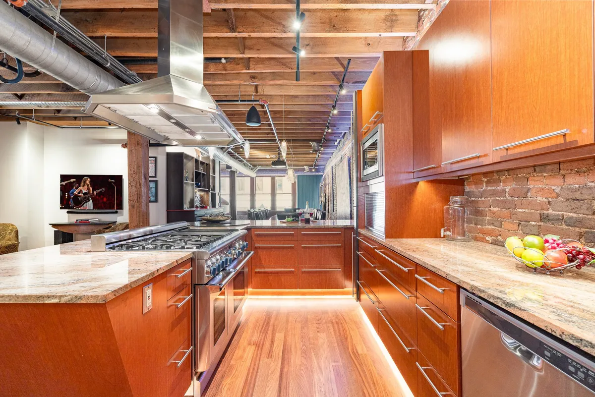A spacious kitchen with an island topped by a granite countertop, state-of-the-art stainless steel appliances, exposed brick wall, and rustic wooden beams overhead. The warm lighting and open layout create a welcoming atmosphere ideal for group cooking or entertaining.