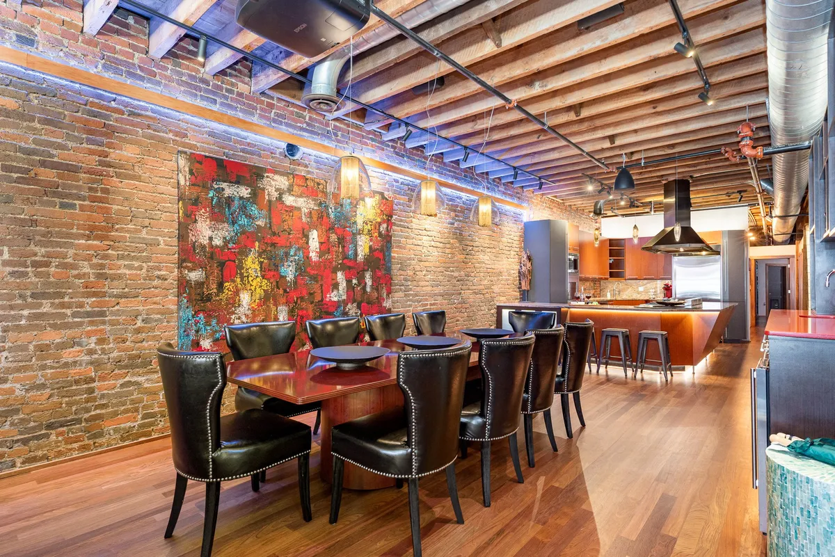 A view from the dining area featuring a long dining table with black chairs, colorful abstract wall art, exposed brick walls, and the kitchen visible in the background. The setup is inviting for large groups and social events.