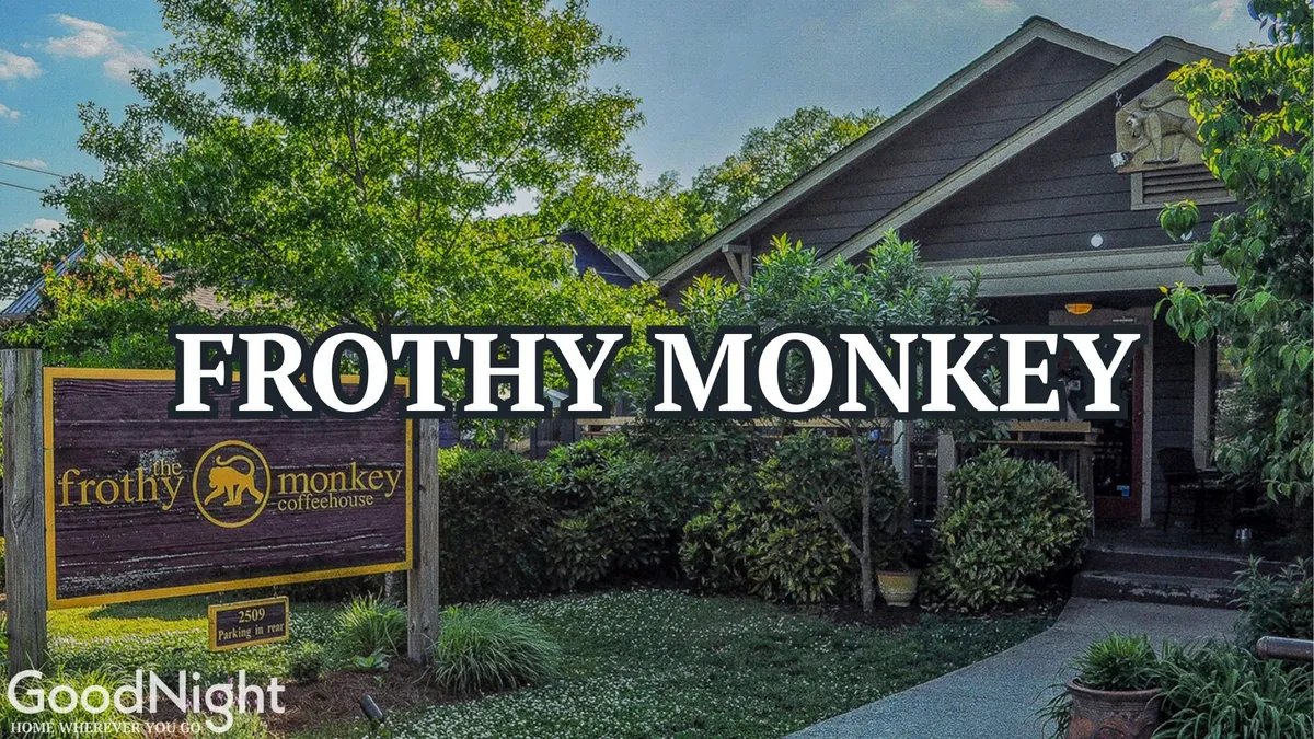 The image captures the welcoming Frothy Monkey coffeehouse entrance surrounded by lush greenery, inviting passersby to stop in for a coffee or light meal. The coffeehouse's prominent sign and the vibrant atmosphere hint at its popularity as a local favorite, with the entrance promising a lively experience inside.