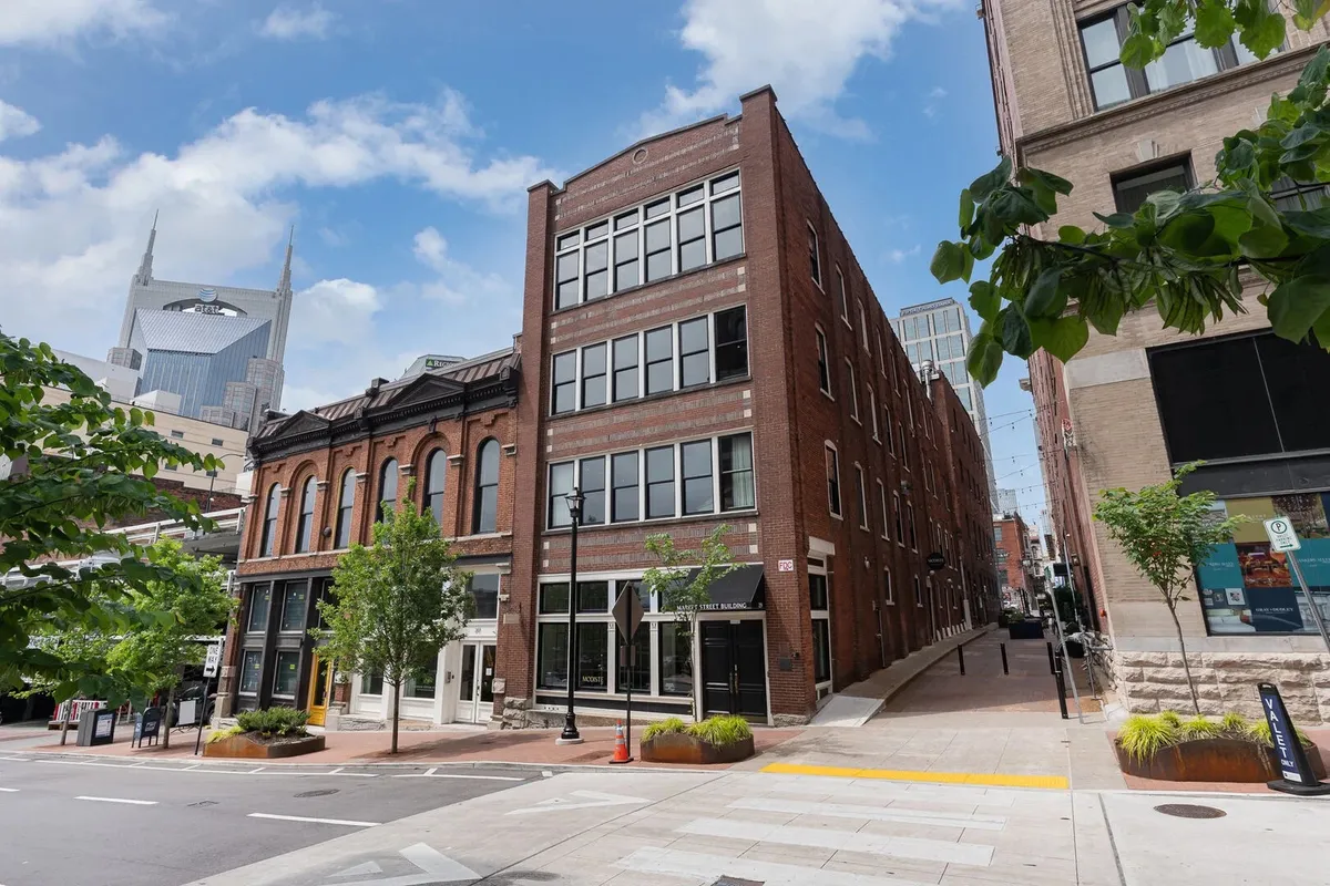 A historic brick building stands prominently in the foreground, flanked by modern skyscrapers and surrounded by green trees and plants. Pedestrians are seen walking and exploring, hinting at the lively and vibrant atmosphere of Nashville. Outdoor dining areas and nearby shops suggest a dynamic urban environment.