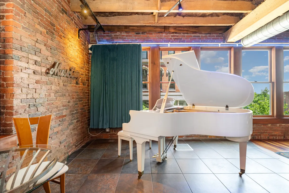 This image shows a grand piano prominently placed in a spacious living area with a backdrop of exposed brick walls and large windows. The room is accented with musical decor, offering a creative and inviting atmosphere.