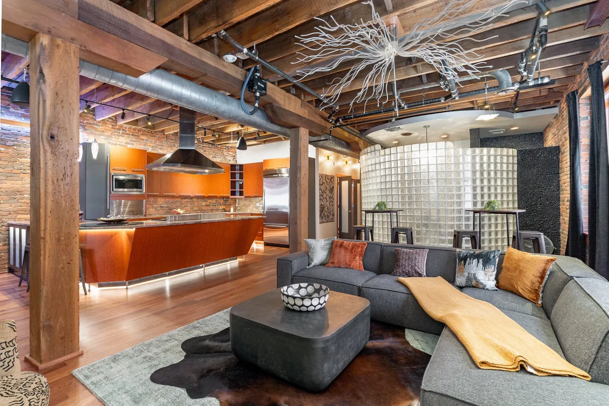 A vibrant interior with a gray sectional sofa, bold orange kitchen cabinetry, a glass block wall, and an artistic ceiling light fixture. The open-plan layout hints at a modern, welcoming entry or living space.