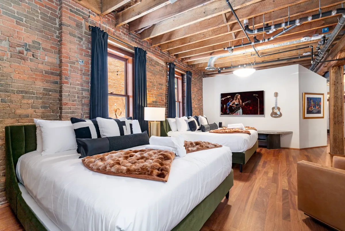 This image displays a spacious bedroom featuring two large beds with plush bedding, wooden beams overhead, exposed brick walls, and a decorative guitar accent. Artistic touches and ample natural light create an inviting and stylish atmosphere ideal for relaxation.